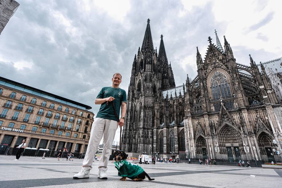 An einem bewölkten Tag steht ein Mann mit einem kleinen Hund vor dem Kölner Dom. Der Hund trägt ein grünes Werder-Trikot, und im Hintergrund ragt der Dom empor.