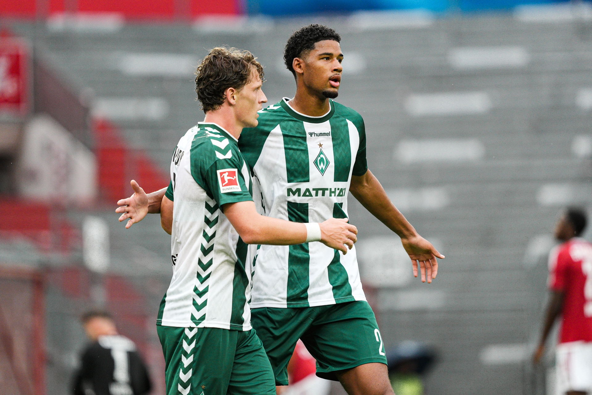 Zwei Fußballspieler in grün-weißen Trikots von Werder Bremen feiern auf dem Spielfeld, im Hintergrund sind verschwommene Zuschauer zu sehen.