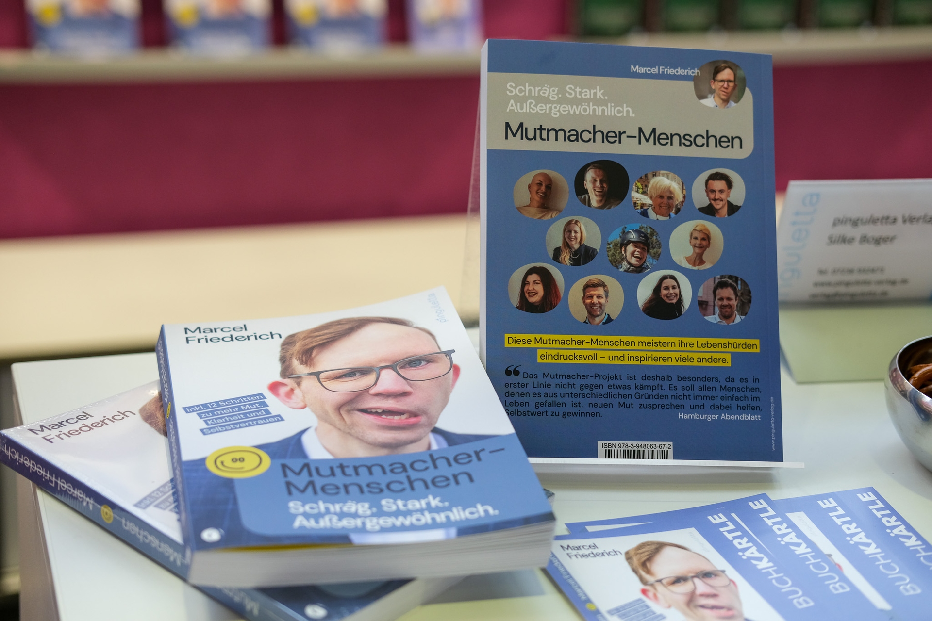 Books titled "Mutmacher-Menschen" on a display, featuring various portraits on the cover against a pink background.