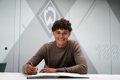 Eine lächelnde Person mit lockigem Haar unterschreibt an einem Tisch ein Dokument, vor einem modernen geometrischen Hintergrund.