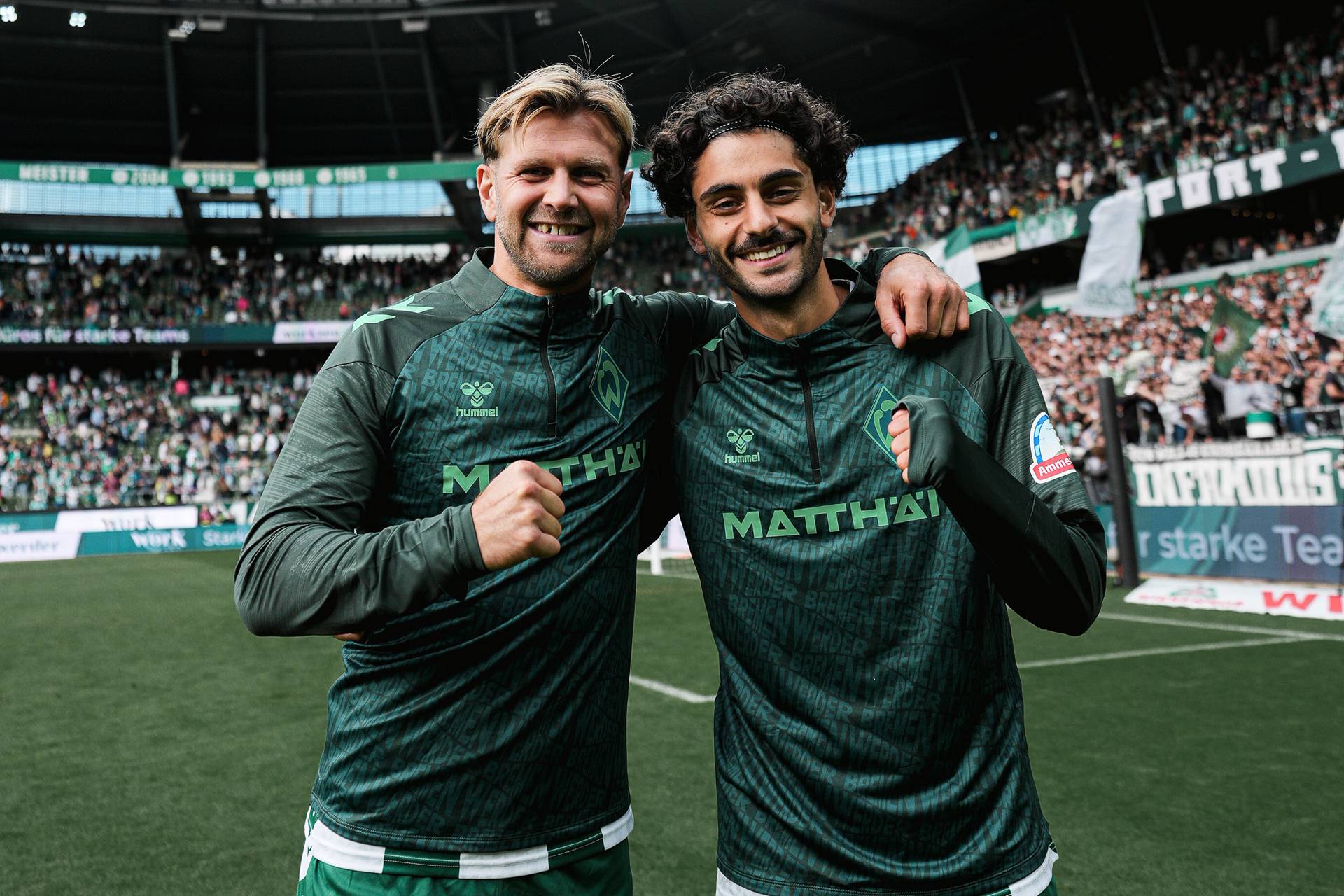 Zwei Fußballspieler in grünen Trikots, die lächelnd Arm in Arm auf einem Spielfeld stehen, im Hintergrund eine jubelnde Zuschauermenge im Stadion.