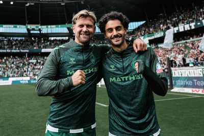 Zwei Fußballspieler in grünen Trikots, die lächelnd Arm in Arm auf einem Spielfeld stehen, im Hintergrund eine jubelnde Zuschauermenge im Stadion.
