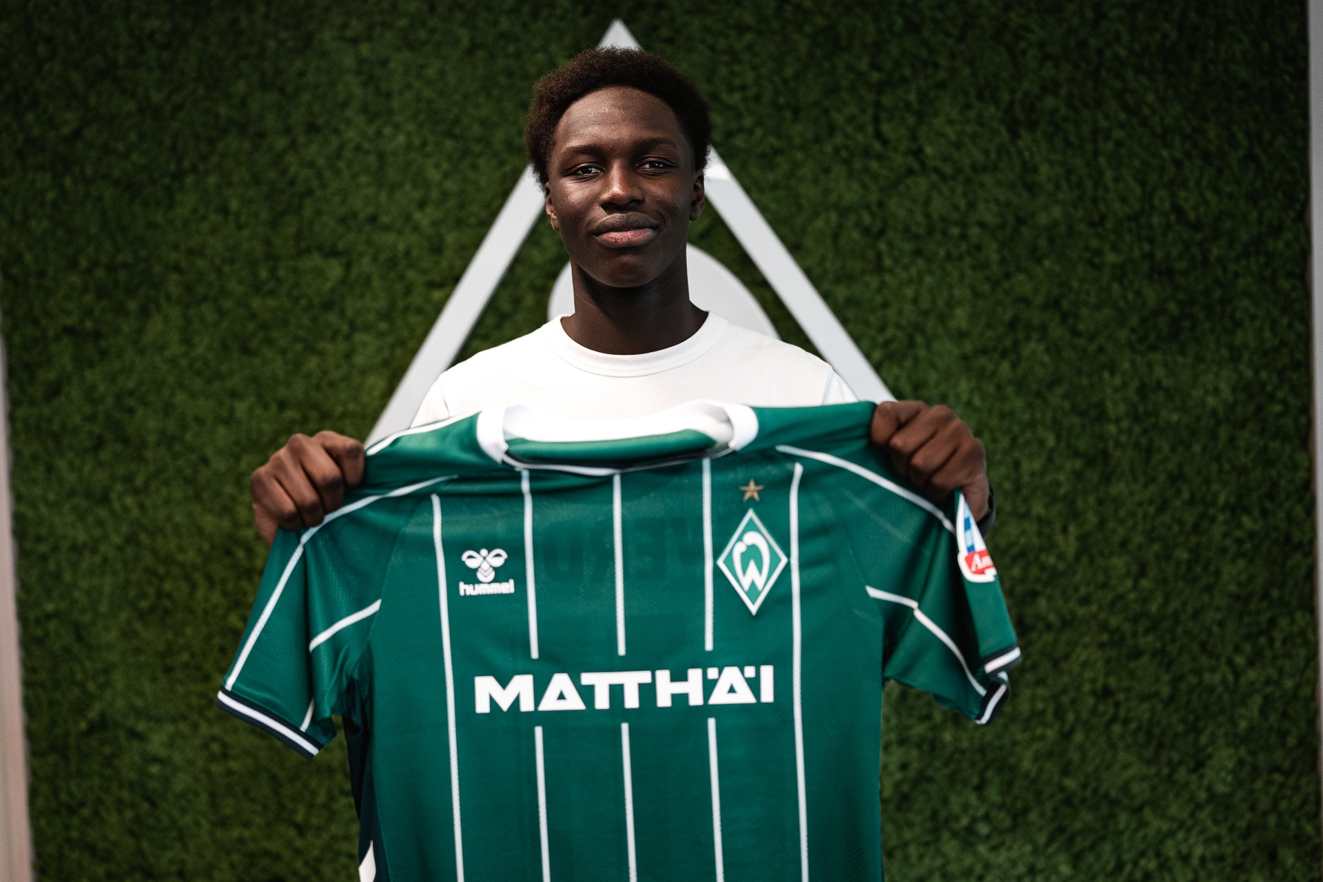 Ramiz Hamouda hält ein Trikot in der Hand und steht vor einer Werder-Raute.