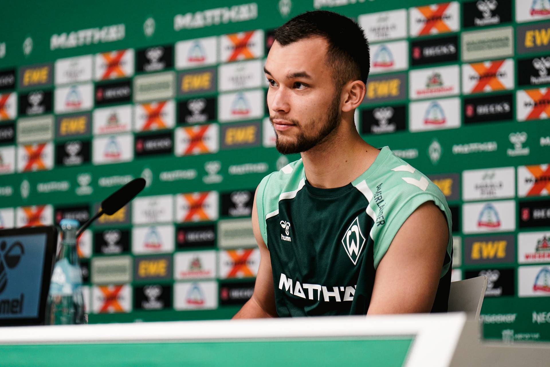 Mio Backhaus an einem Pressekonferenztisch vor einem Hintergrund mit dem Logo seines Teams, bekleidet mit einem grünen Trikot, auf dem Logos und der Schriftzug „MATTHÄI“ zu sehen sind.
