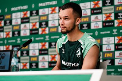 Mio Backhaus an einem Pressekonferenztisch vor einem Hintergrund mit dem Logo seines Teams, bekleidet mit einem grünen Trikot, auf dem Logos und der Schriftzug „MATTHÄI“ zu sehen sind.