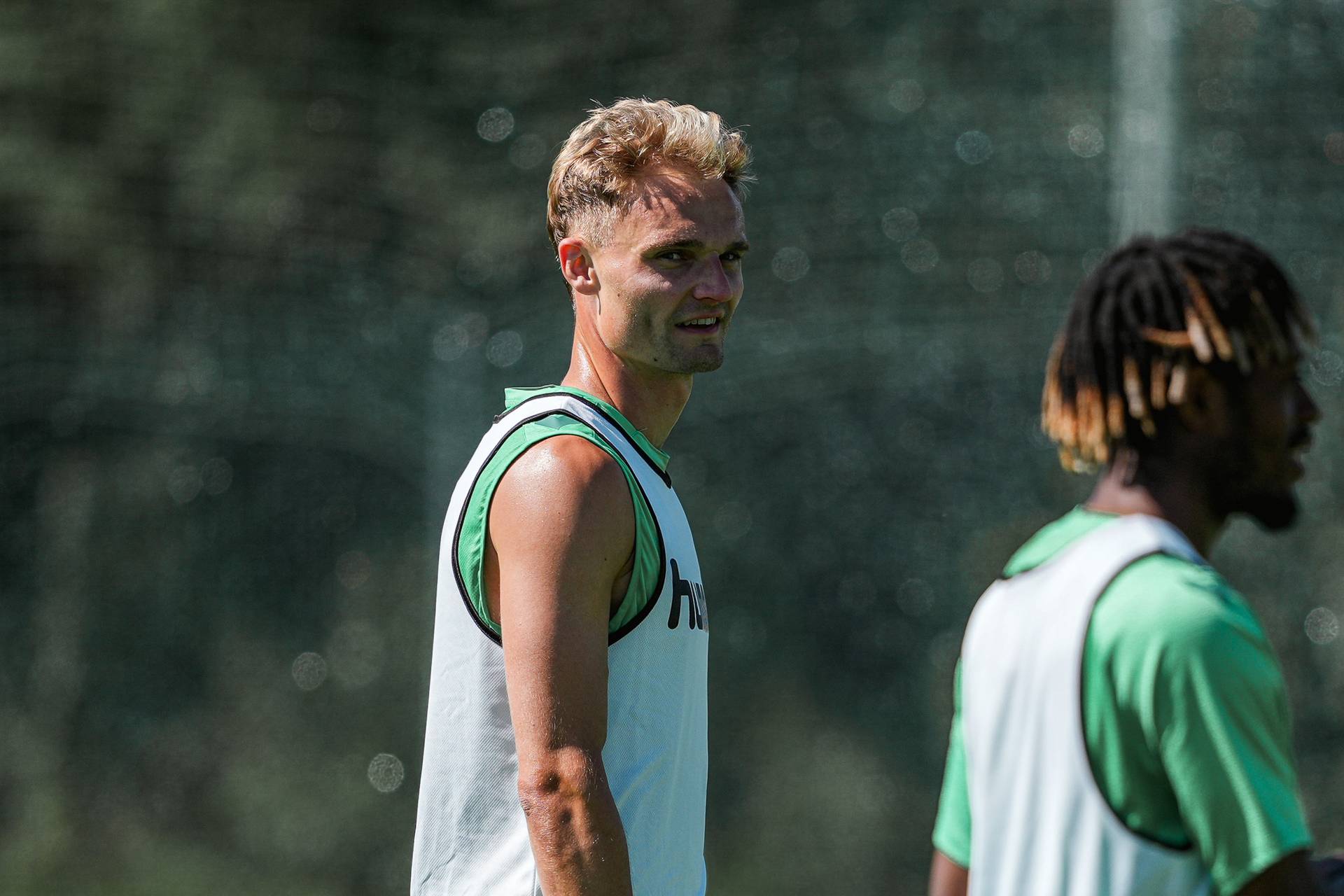 Zwei Sportler in ärmelloser Sportbekleidung beim Training im Freien; einer blickt in die Kamera, während sich das Sonnenlicht auf seiner Haut spiegelt.