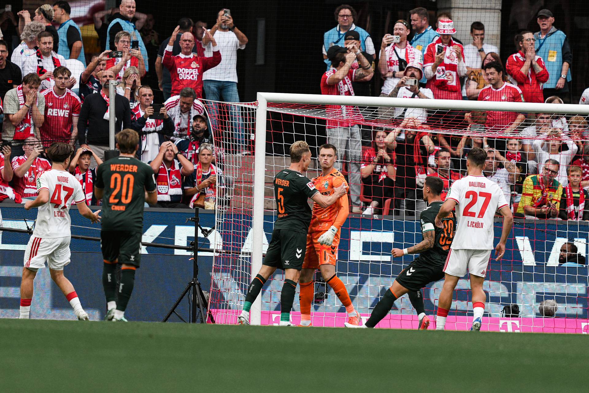 Während eines Spiels versammeln sich die Fußballspieler in der Nähe des Torpfostens, während im Hintergrund Fans in roten Trikots jubeln und mitfiebern.