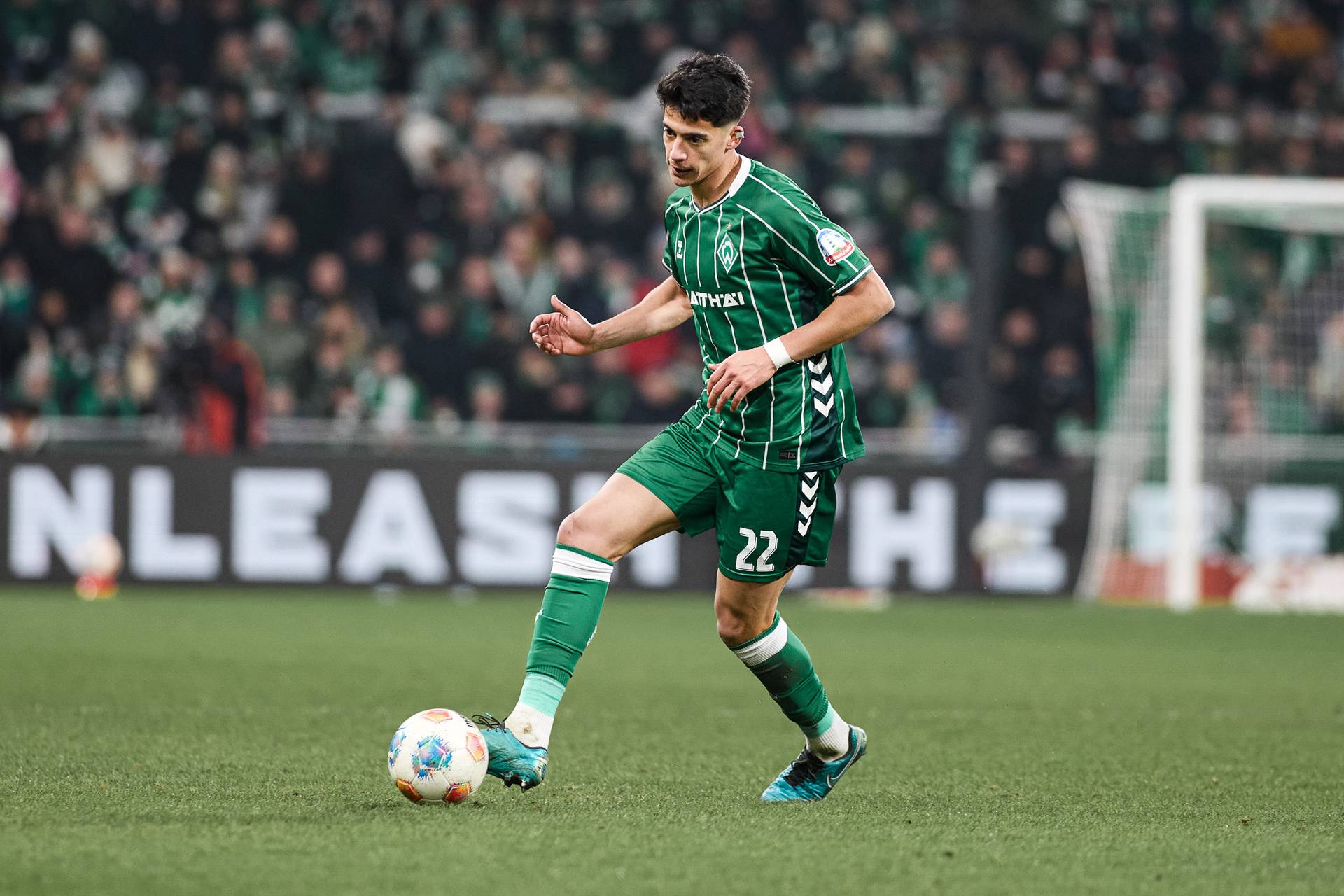 Ein Fußballspieler in grün-weißem Trikot dribbelt den Ball auf dem Spielfeld, im Hintergrund sind eine verschwommene Menschenmenge und ein Torpfosten zu sehen.
