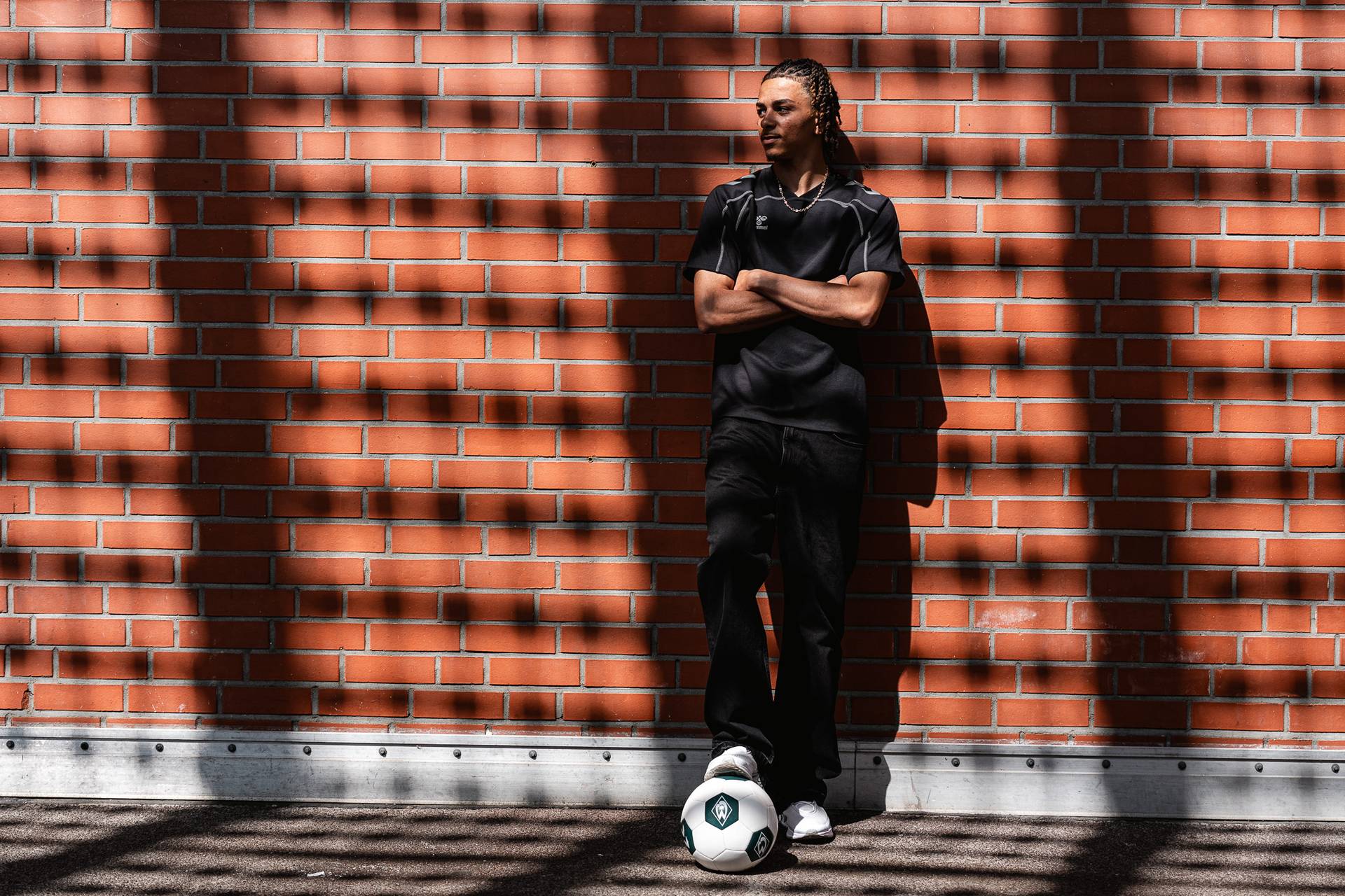 Kenny Quetant in black clothing leans against a sunlit brick wall with his arms crossed, a football lying by his feet.