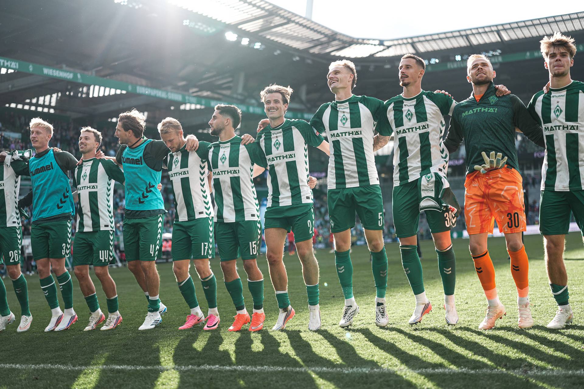 Eine Fußballmannschaft in grün-weißen Trikots, die sich unterm Arm gegriffen hat und auf dem Spielfeld in einem sonnenbeschienenen Stadion jubelt.