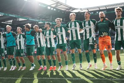 Eine Fußballmannschaft in grün-weißen Trikots, die sich unterm Arm gegriffen hat und auf dem Spielfeld in einem sonnenbeschienenen Stadion jubelt.