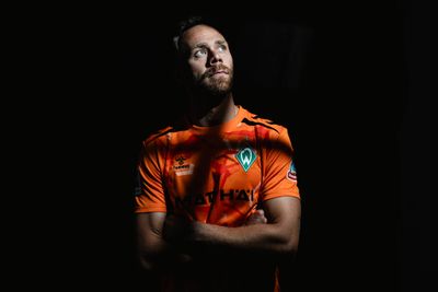 Fußballspieler im orangefarbenen Trikot mit grünem Logo, die Arme verschränkt, blickt vor einem dunklen Hintergrund in dramatischer Beleuchtung nach oben.