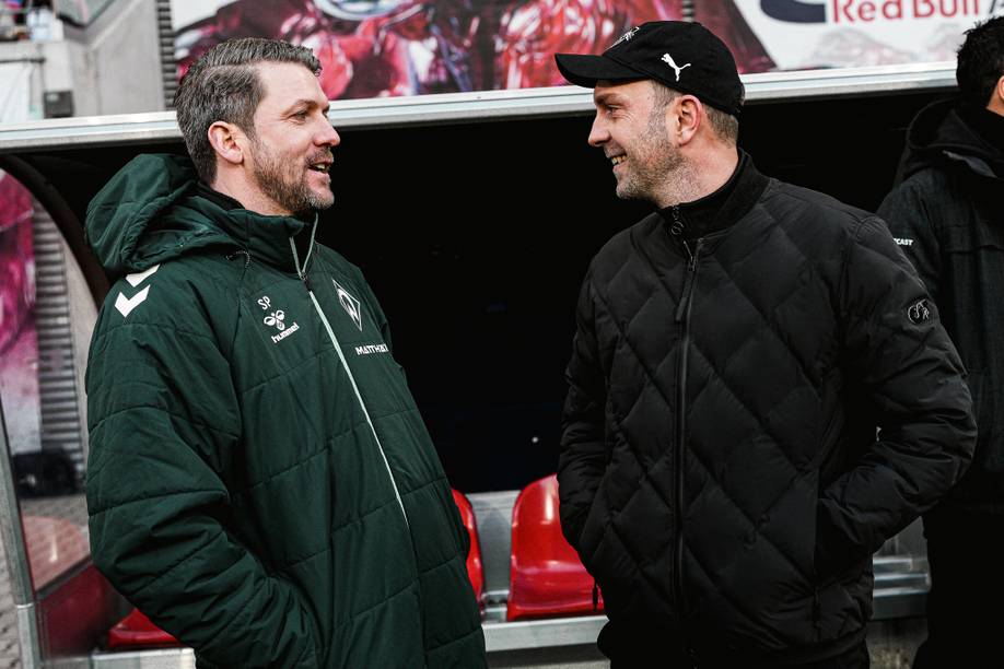 Zwei Männer in Winterjacken und Mützen lächeln einander an, während sie in einem Stadionbereich mit roten Stühlen und einer Werbetafel im Hintergrund stehen.
