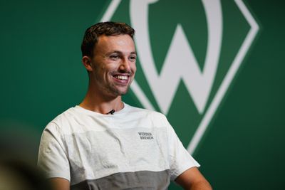 Ein lächelnder Mann in einem weißen T-Shirt mit der Aufschrift „Werder Bremen“, der vor einem grünen Hintergrund mit einem großen Logo sitzt.
