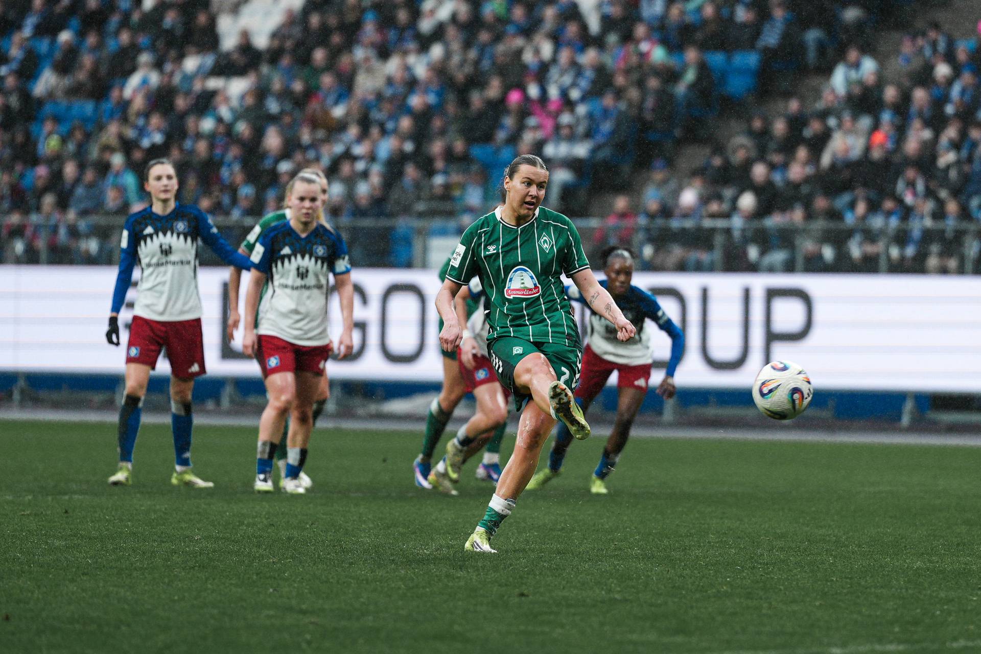 Ein Fußballspieler in einem grünen Trikot schießt während eines Spiels den Ball, im Hintergrund sind seine Teamkollegen und Gegner zu sehen, und auf den Tribünen sitzt eine Menschenmenge.