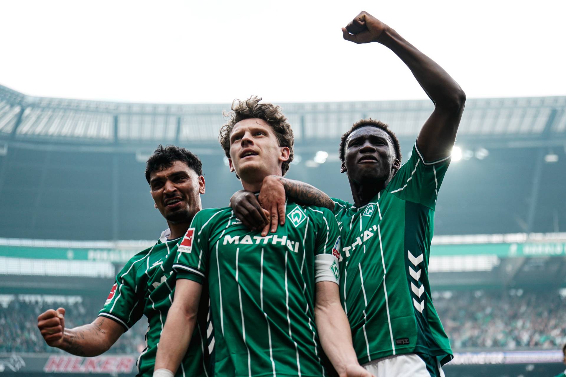 Drei Fußballspieler in grünen Trikots feiern, einer reckt die Faust in die Höhe, in einem Stadion vor jubelnden Zuschauern.