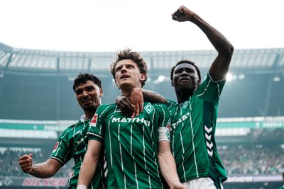 Drei Fußballspieler in grünen Trikots feiern, einer reckt die Faust in die Höhe, in einem Stadion vor jubelnden Zuschauern.