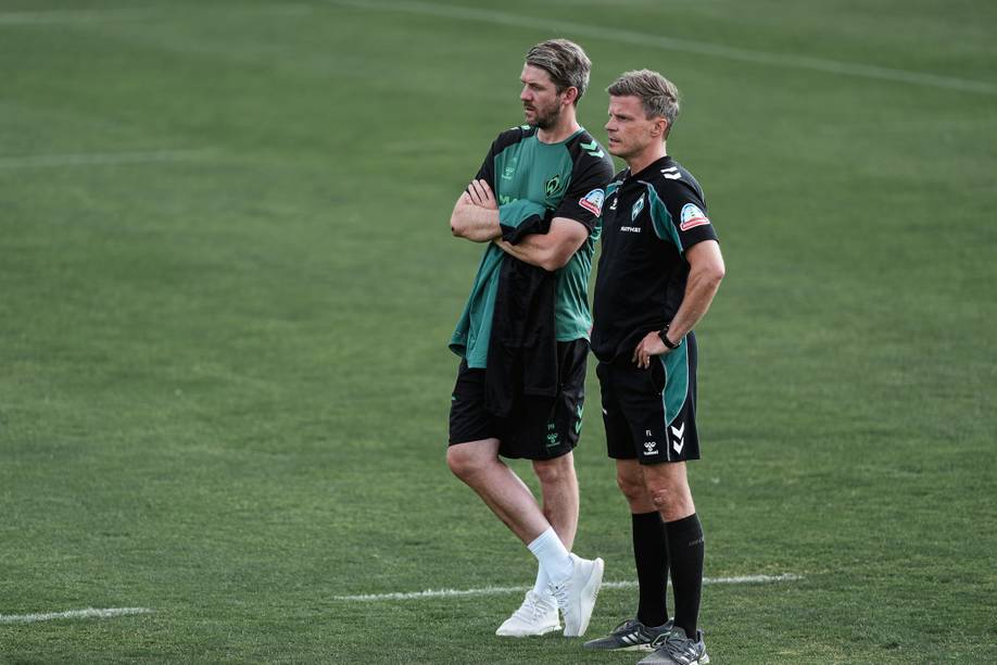 Zwei Männer in Sportkleidung stehen auf einer Wiese und blicken konzentriert auf etwas außerhalb des Bildausschnitts. Der eine hat die Arme verschränkt, der andere die Hände in die Hüften gestemmt.