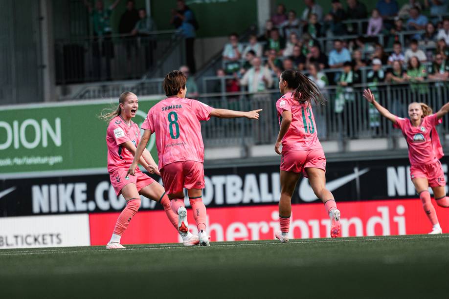 Fußballspieler in rosa Trikots feiern auf dem Spielfeld ein Tor, umgeben von jubelnden Zuschauern im Stadion.