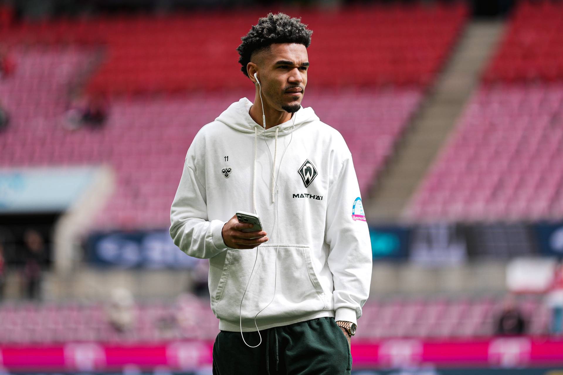 Eine Person in einem weißen Kapuzenpulli mit Logos, die über Kopfhörer Musik hört, hält ein Handy in der Hand, während sie in einem Stadion mit roten Sitzplätzen steht.