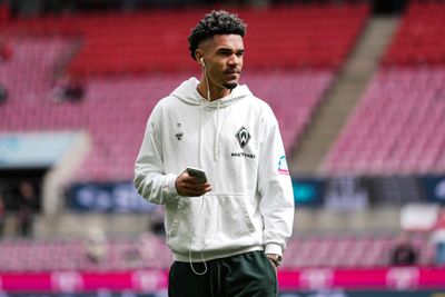 Eine Person in einem weißen Kapuzenpulli mit Logos, die über Kopfhörer Musik hört, hält ein Handy in der Hand, während sie in einem Stadion mit roten Sitzplätzen steht.