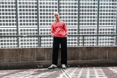 Eine Person in einem roten Pullover und einer schwarzen Hose steht lässig mit den Händen in den Taschen vor einer Betonwand vor einem Hintergrund mit Gittermuster.