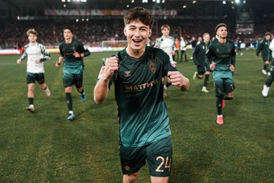 Ein Fußballspieler in einem grün-schwarzen Trikot jubelt freudig, während seine Teamkollegen hinter ihm auf dem Stadionfeld laufen.