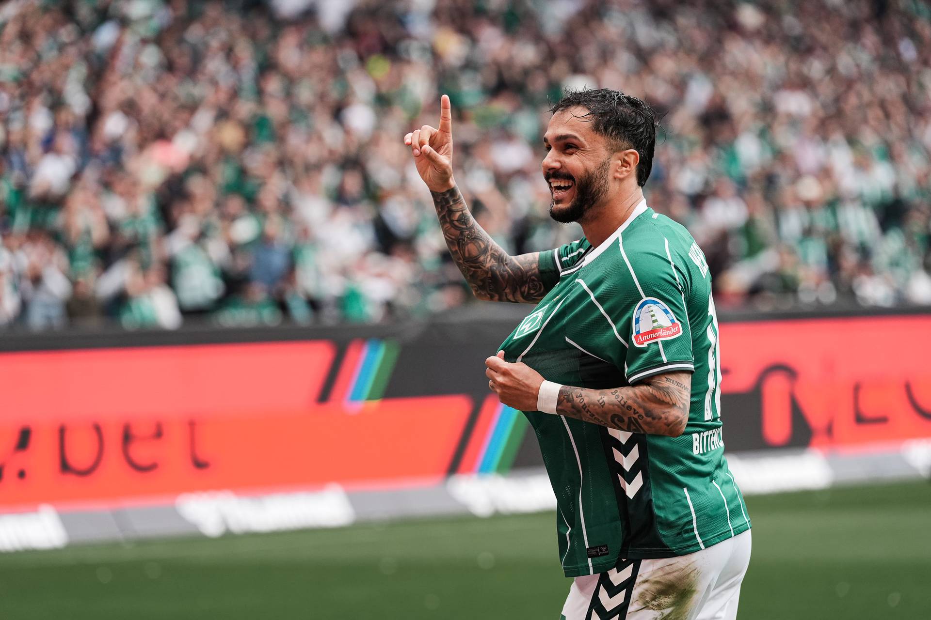 Leonardo Bittencourt showing off the Werder badge on his shirt and holding one finger in the air.