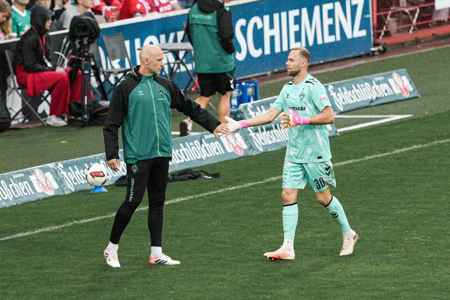 Ein Fußballtrainer und ein Torwart in einheitlicher grüner Kleidung geben sich während eines Spiels an der Seitenlinie die Hand, im Hintergrund sind Zuschauer zu sehen.