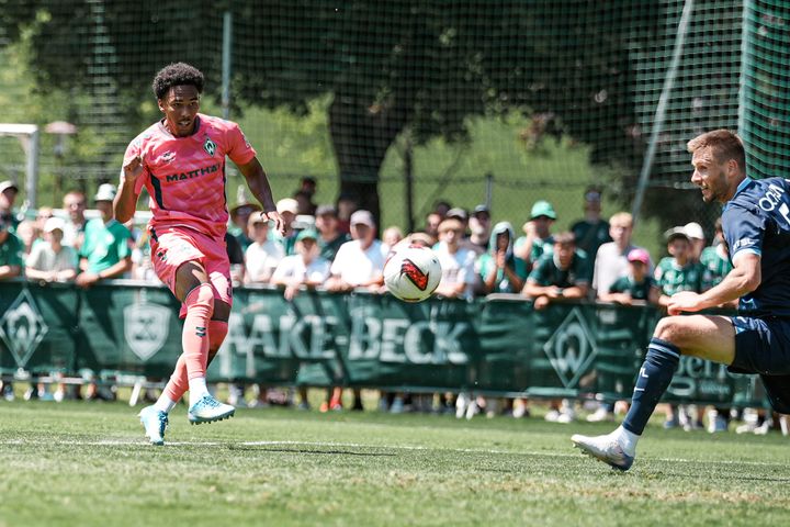 Ein Fußballspieler im rosa Trikot versucht einen Schuss, während ein anderer Spieler in Blau auf dem sonnigen Spielfeld in einen Zweikampf geht, um den Ball abzufangen; im Hintergrund beobachten Zuschauer das Geschehen.