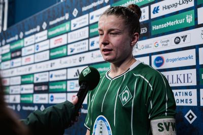 Fußballspieler in einem grünen Trikot wird mit einem Mikrofon interviewt, während er vor einem Sponsoren-Hintergrund steht.