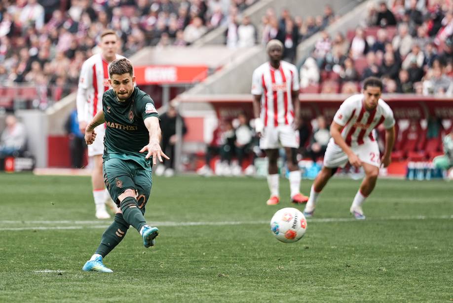 Ein Fußballspieler in grüner Spielkleidung schießt während eines Spiels den Ball, im Hintergrund sind Spieler der rot-weißen Mannschaft und Zuschauer zu sehen.