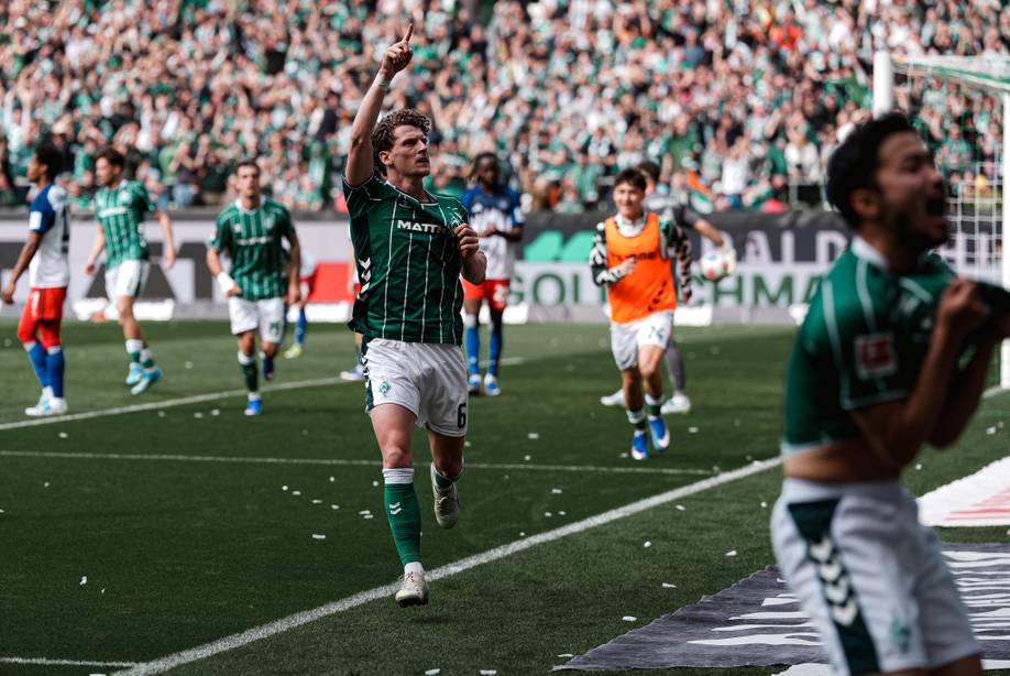 Ein Fußballspieler in grüner Kleidung jubelt auf dem Spielfeld und zeigt nach oben. Im Hintergrund sind seine Mannschaftskameraden und die Zuschauer zu sehen, die für eine lebhafte Atmosphäre sorgen.