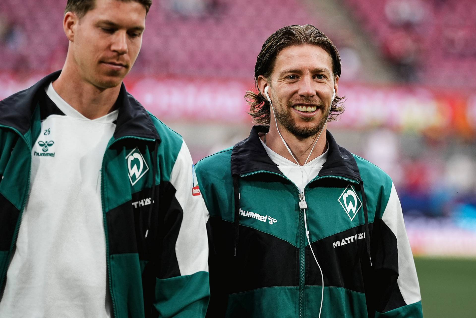 Zwei Fußballspieler in grünen Trainingsanzügen, von denen einer lächelt und Kopfhörer trägt, stehen auf einem Spielfeld vor einem verschwommenen Stadionhintergrund.