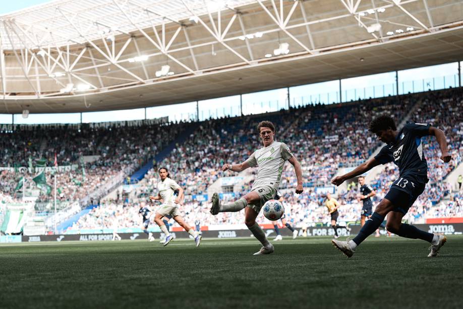 Zwei Fußballspieler in Aktion auf einem sonnenbeschienenen Stadionrasen, im Hintergrund jubelt das vollbesetzte Publikum.