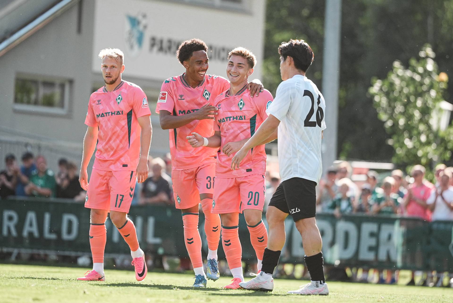 Vier Fußballspieler, drei in rosa Trikots und einer in einem weißen, stehen auf einem sonnigen Spielfeld. Zwei der Spieler in rosa Trikots lächeln und jubeln.