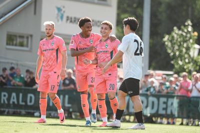 Vier Fußballspieler, drei in rosa Trikots und einer in einem weißen, stehen auf einem sonnigen Spielfeld. Zwei der Spieler in rosa Trikots lächeln und jubeln.