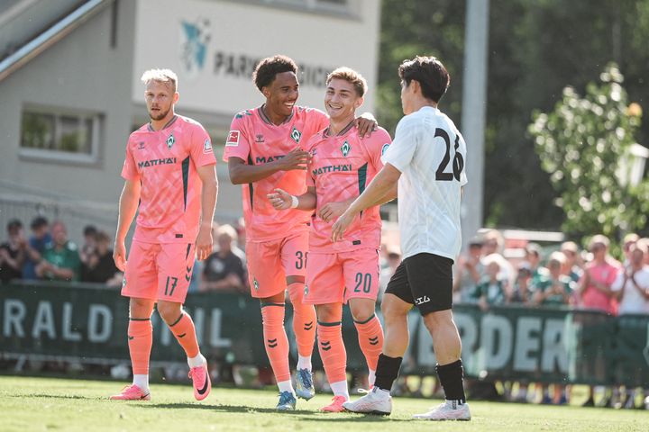 Vier Fußballspieler, drei in rosa Trikots und einer in einem weißen, stehen auf einem sonnigen Spielfeld. Zwei der Spieler in rosa Trikots lächeln und jubeln.