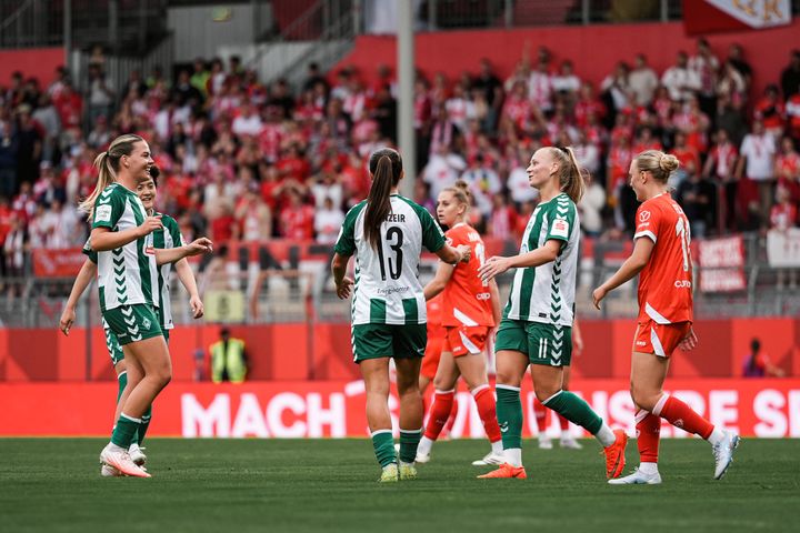 Fußballerinnen in grün-weißen Trikots feiern auf dem Spielfeld, im Hintergrund sind Fans in roten Trikots zu sehen.