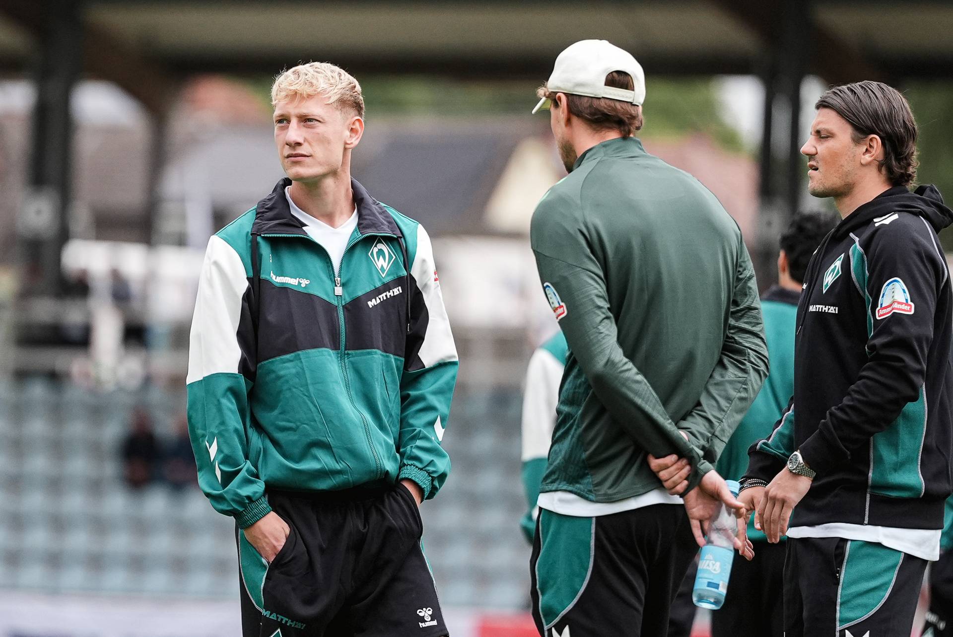 Drei Männer in grüner und schwarzer Sportkleidung unterhalten sich am Spielfeldrand eines Stadions; einer von ihnen hält eine Wasserflasche in der Hand.
