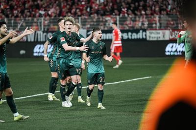 Fußballspieler in blaugrünen Trikots feiern ein Tor auf dem Spielfeld; im Hintergrund rot gekleidete Fans.