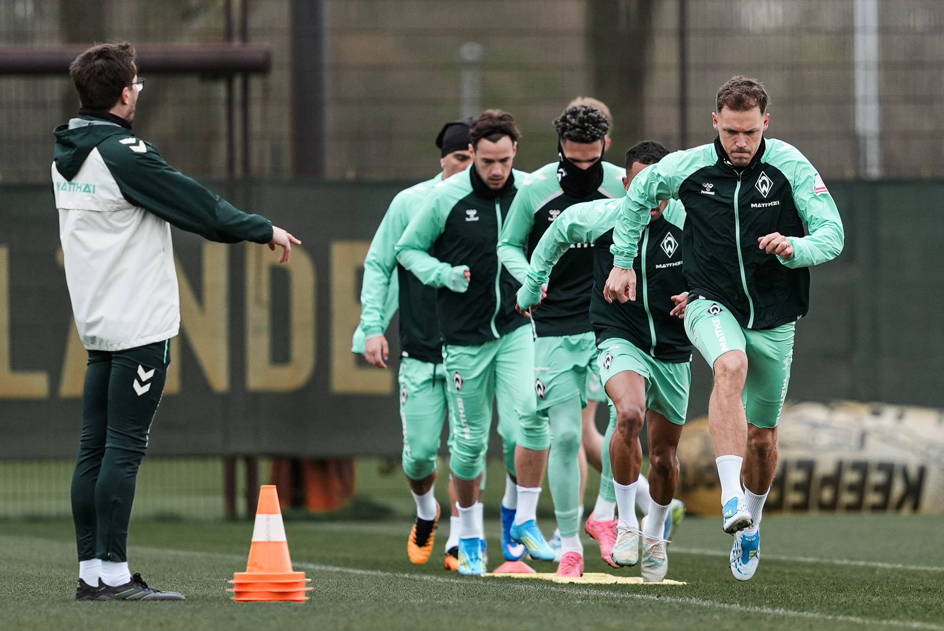 Fußballspieler in grüner Trainingskleidung joggen in einer Reihe über den Platz und folgen einem Trainer, der sich in der Nähe orangefarbener Kegel befindet.