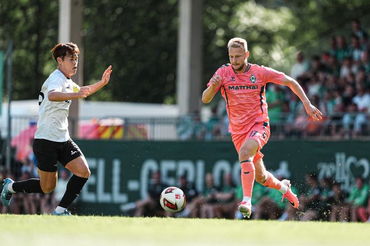Zwei Fußballspieler in Aktion: Ein Spieler in Weiß verteidigt gegen einen Spieler in Rosa, während sie auf einem sonnigen Spielfeld dem Ball nachjagen.