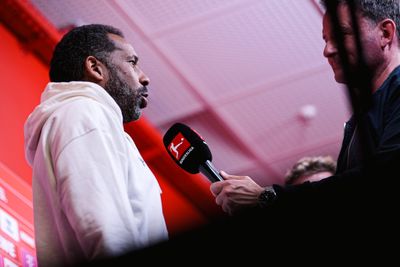 Ein Mann in einem weißen Kapuzenpulli spricht vor einem rot beleuchteten Hintergrund in ein Bundesliga-Mikrofon, das von einer anderen Person gehalten wird.