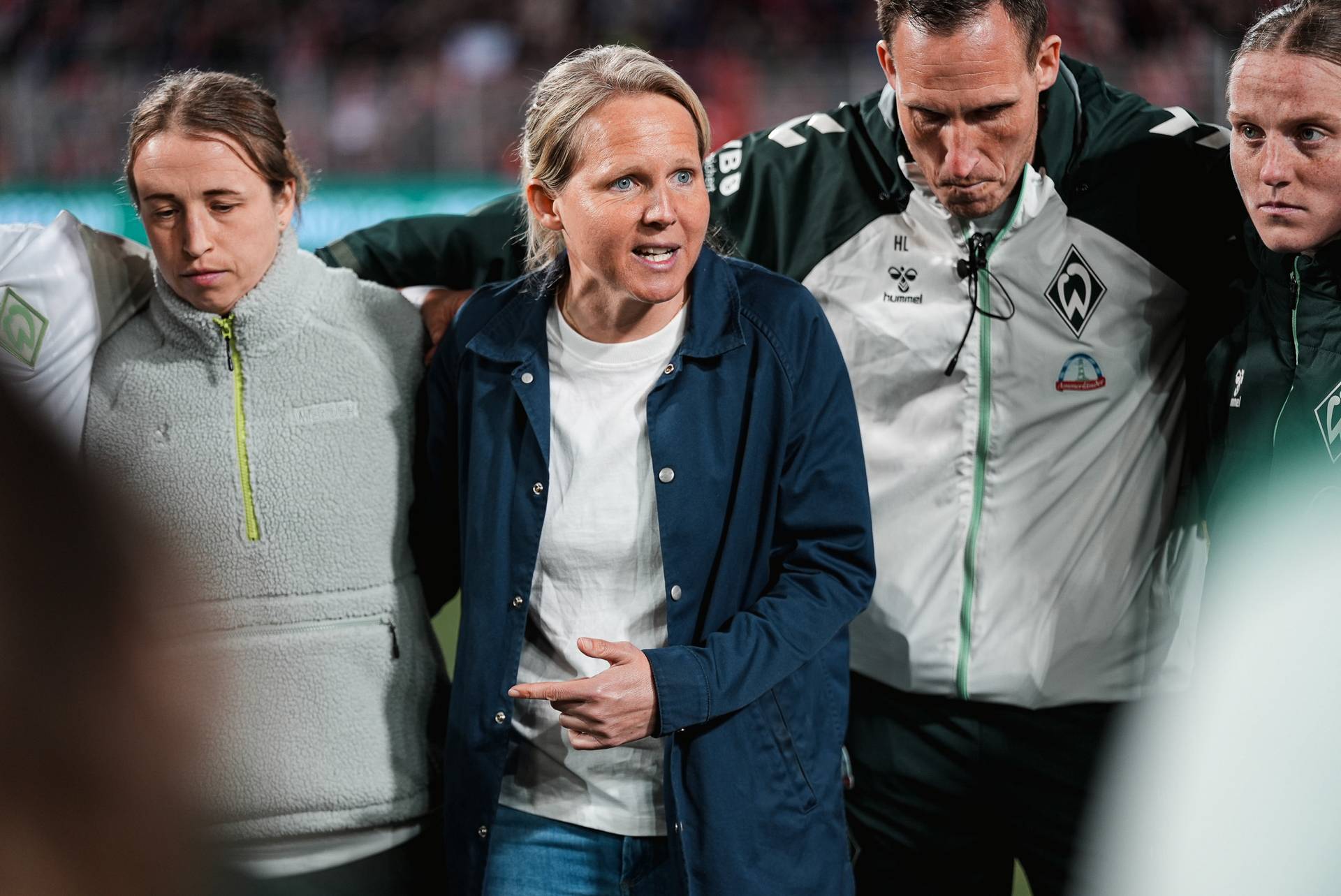 Eine Gruppe von Fußballspielerinnen und eine Trainerin stehen in einer Besprechungsrunde auf dem Spielfeld und unterhalten sich; sie tragen ihre Mannschaftsjacken.