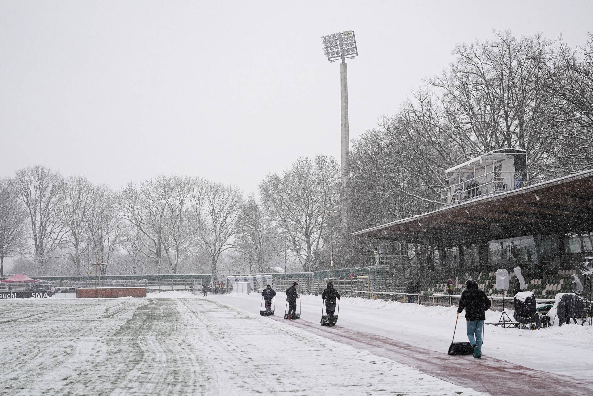 Menschen räumen mit Schaufeln Schnee von einem Sportplatz. Der Boden ist mit Schnee bedeckt, im Hintergrund stehen kahle Bäume. Ein hoher Lichtmast ist zu sehen.