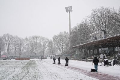 Menschen räumen mit Schaufeln Schnee von einem Sportplatz. Der Boden ist mit Schnee bedeckt, im Hintergrund stehen kahle Bäume. Ein hoher Lichtmast ist zu sehen.