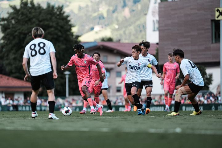 Fußballspieler in rosa-weißen Trikots bestreiten ein Spiel auf einem grünen Rasen, im Hintergrund sind Bäume und Gebäude zu sehen.