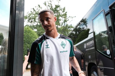 Ein Mann in Sportkleidung steht vor einem Bus und trägt Kopfhörer. Im Hintergrund sind Bäume und ein Bus mit der Aufschrift „Jacobs“ zu sehen.