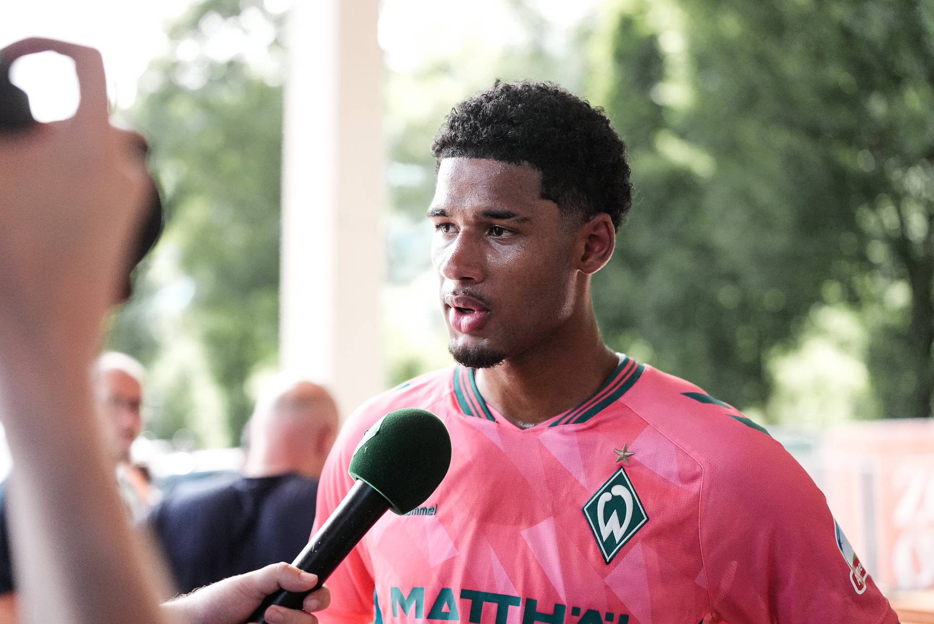 Ein Fußballspieler in einem rosa Trikot von Werder Bremen wird interviewt, während er ein grünes Mikrofon in der Hand hält; im Hintergrund sind verschwommen Bäume und Menschen zu sehen.